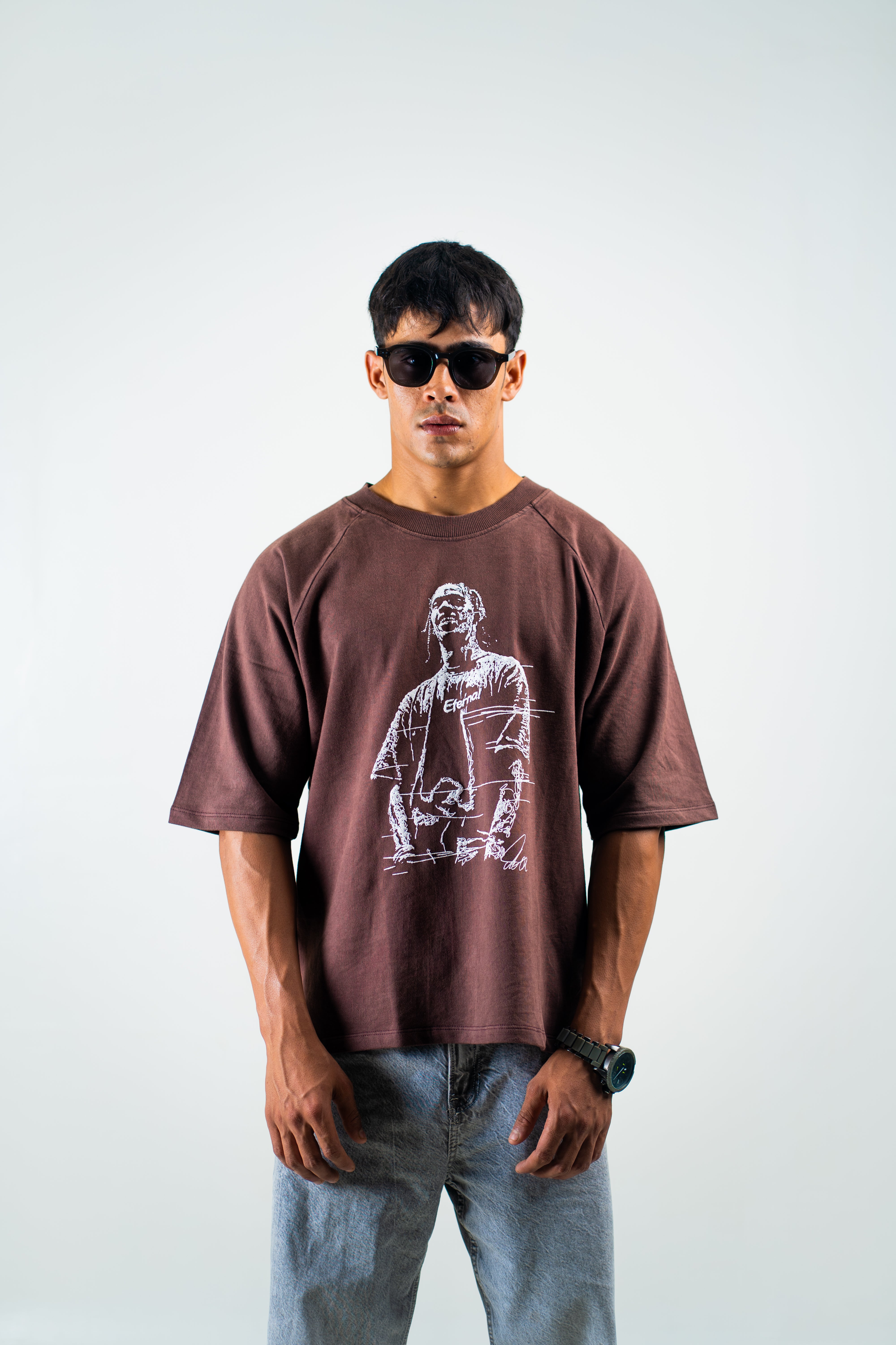 A man wearing a brown oversized graphic t-shirt with a white sketch-style print of a man, paired with light blue jeans and black sunglasses, standing against a light gray background.